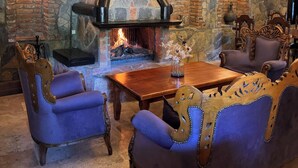 A cozy interior with a stone fireplace, wooden beams, and blue upholstered chairs around a wooden table. - Manastır Mandarin (Tahtakuslar)