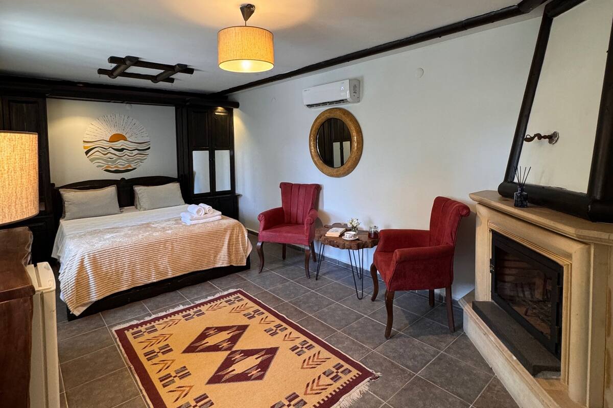 Una habitación de hotel con cama, un sillón rojo, una mesita, chimenea y una lámpara en el techo.