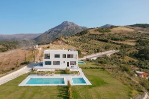 A modern two-story building with a pool and outdoor seating, surrounded by hilly terrain and a mountain in the background.