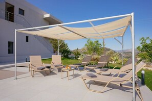 An outdoor seating area with lounge chairs and a table, shaded by a canopy, in front of a modern building with a clear view of the surrounding landscape. - Luxury Villa in Crete with 3 Pools, Stunning Views & Total Privacy (Argiroupoli)