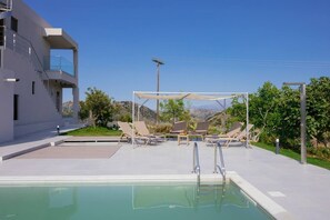 A modern outdoor pool area with lounge chairs and a pergola, surrounded by a landscaped garden and a mountain view.
