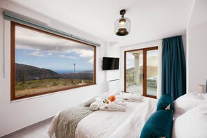 Quarto de hotel com uma janela ampla, cama com lençóis brancos, almofada azul-turquesa e luminária no teto.
