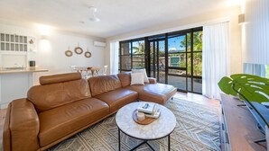 A living room with a brown leather sofa, a round coffee table, and a large window with a view of greenery outside. - Airlie Beach main street 2 minute walk (Airlie Beach)