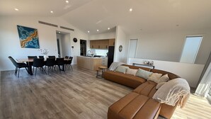Un salon moderne avec un canapé en cuir marron, un coin repas avec une table et des chaises en bois, et une cuisine en arrière-plan.
