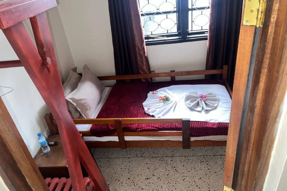 A single bed with a wooden frame, a red bedspread, and a window with a hexagonal pattern.