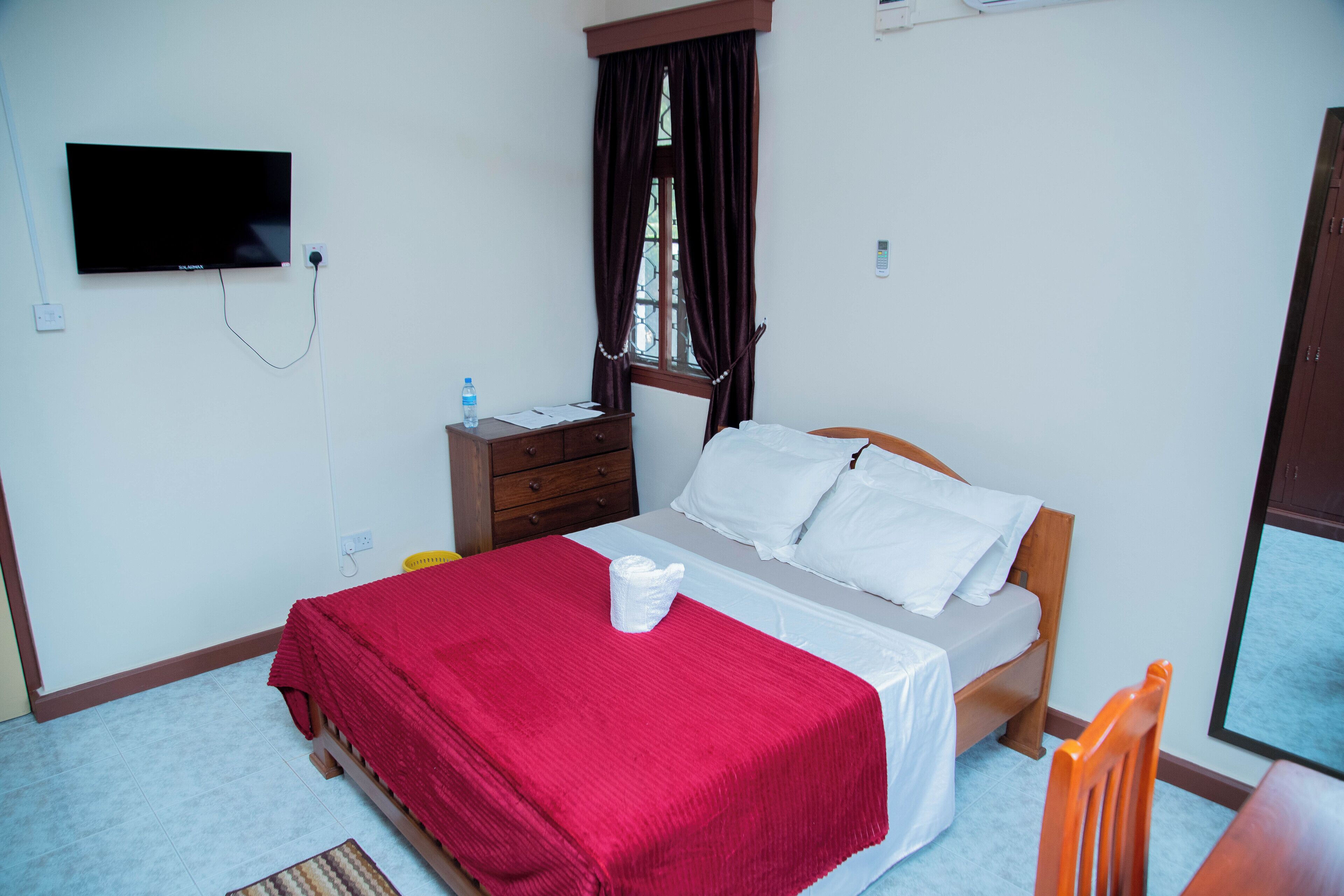 Quarto com cama, televisão fixada na parede, janela com cortinas, mesa de cabeceira de madeira e uma cadeira.