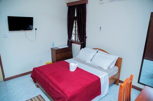 Quarto com cama, televisão fixada na parede, janela com cortinas, mesa de cabeceira de madeira e uma cadeira.