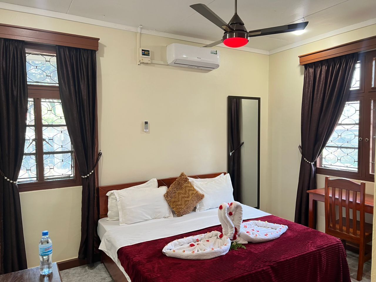 A hotel room with a bed, a ceiling fan, an air conditioner, a water bottle, and a window with curtains.