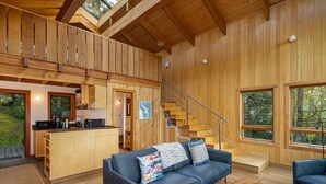 A spacious living area with a wooden interior, a sofa with colorful cushions, a coffee table, and a view of the outdoors through large windows.