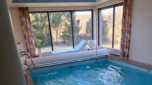 An indoor swimming pool with clear glass walls, a lounge area with white chairs, and a view of trees outside. - Holiday Home 'With Pool Hall' with Private Terrace, Garden and Wi-Fi (Burgthann)