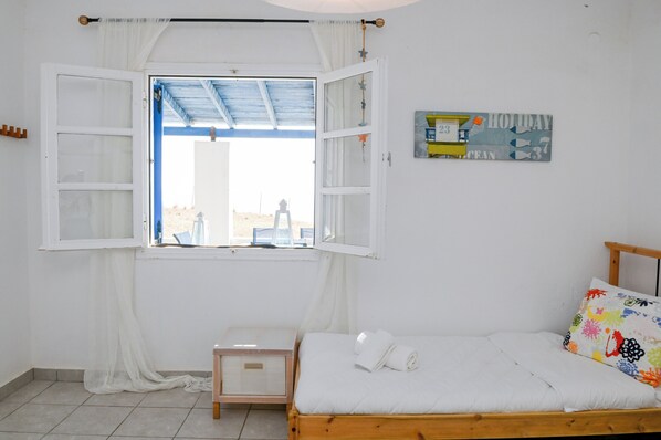 A room with a bed, a small table, a window with curtains, and a wall decoration. - Santorini View House (Santorini)