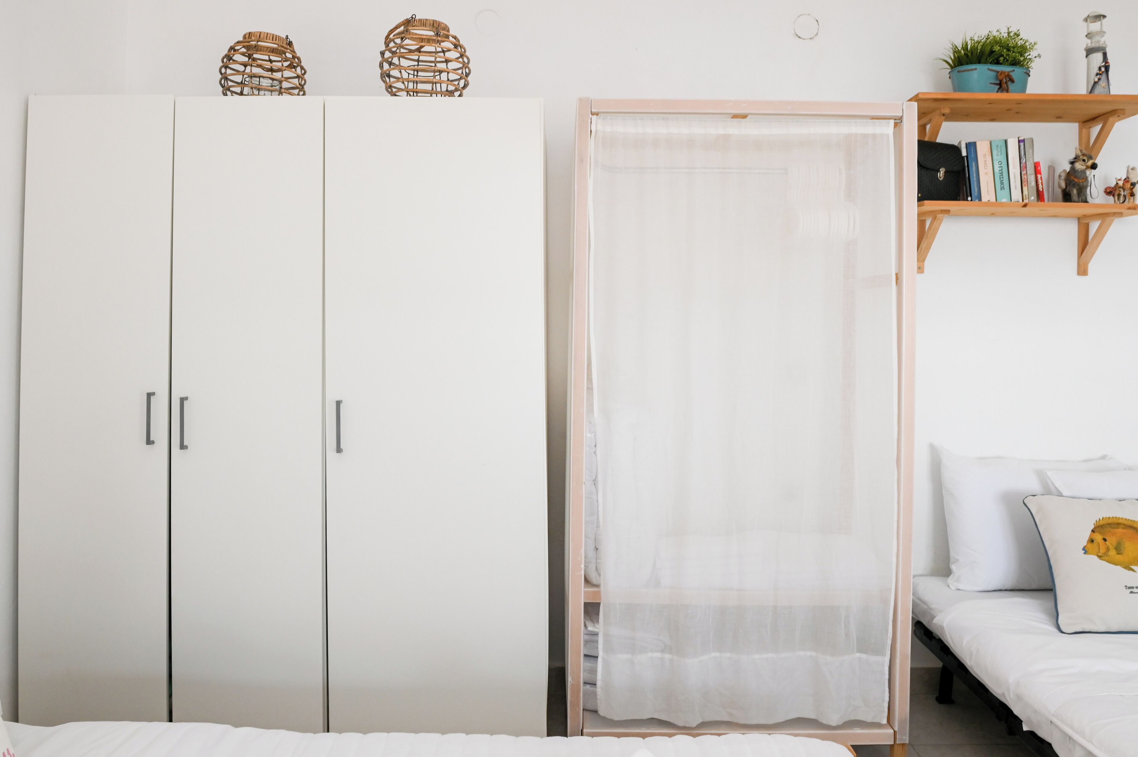 Un dormitorio con cama, armario, mesita de noche, estante con libros y una planta, y ventana con cortina.