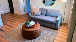 Un salon avec un canapé gris, une table basse ronde ornée d’une plante, un miroir rond accroché au mur et une lampe sur pied.