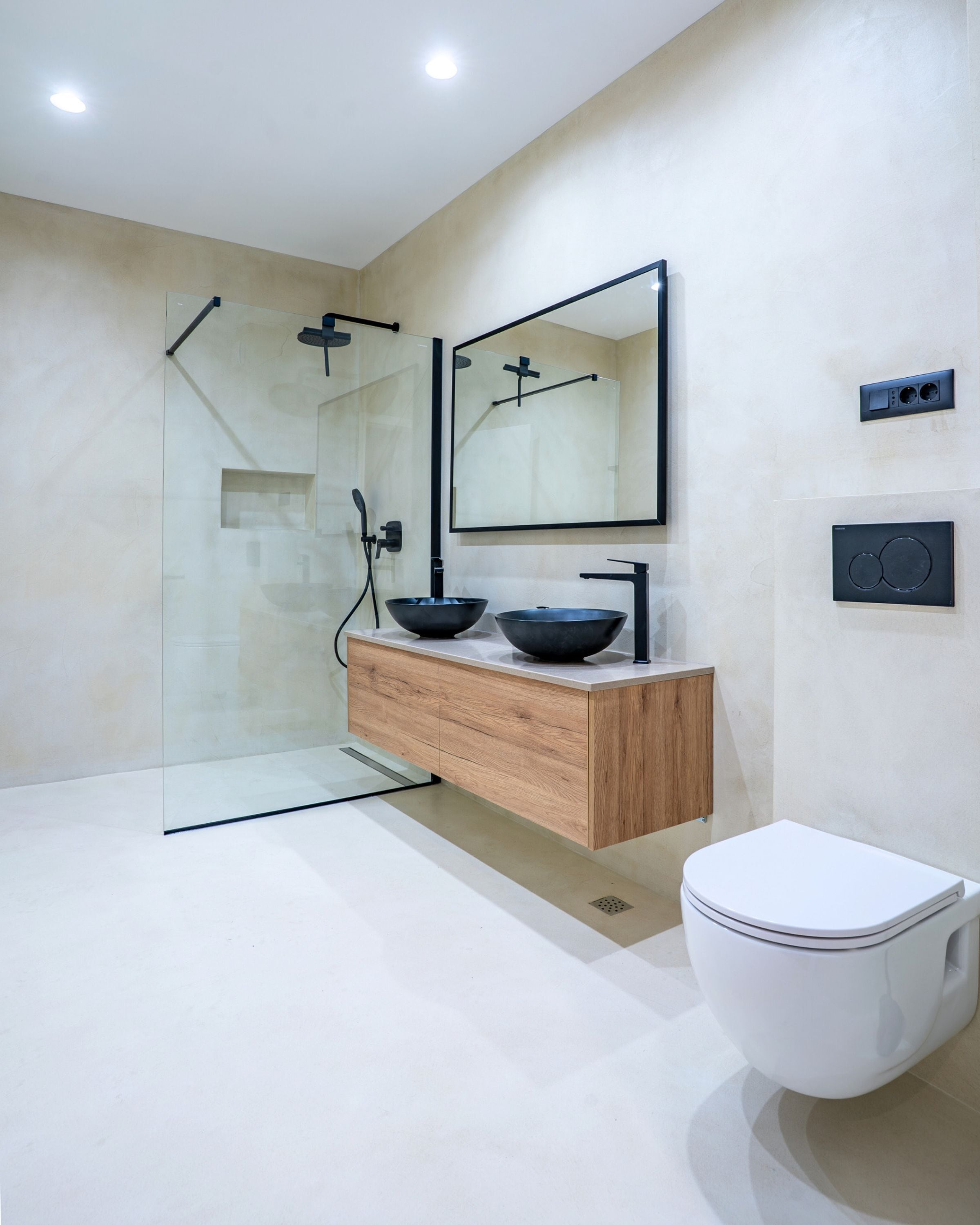 A modern bathroom with a glass shower enclosure, a wooden vanity with two black bowls, a white toilet, and a wall-mounted mirror.