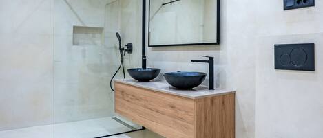 A modern bathroom with a glass shower enclosure, a wooden vanity with two black bowls, a white toilet, and a wall-mounted mirror.