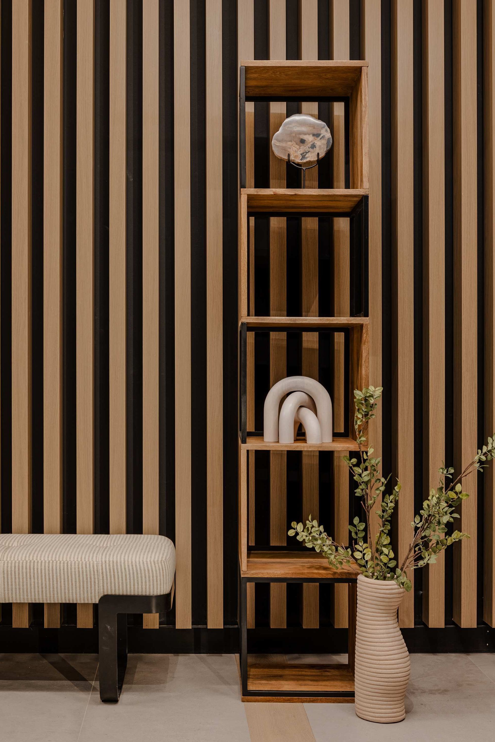 Un interior moderno con una pared de madera a rayas, una alta estantería de madera con objetos decorativos, un banco y un florero con plantas.