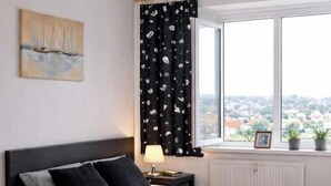 Une chambre à coucher avec un lit, une table de chevet, une lampe, une fenêtre avec des rideaux, une vue sur la ville et une table sur laquelle est posé un vase de fleurs.