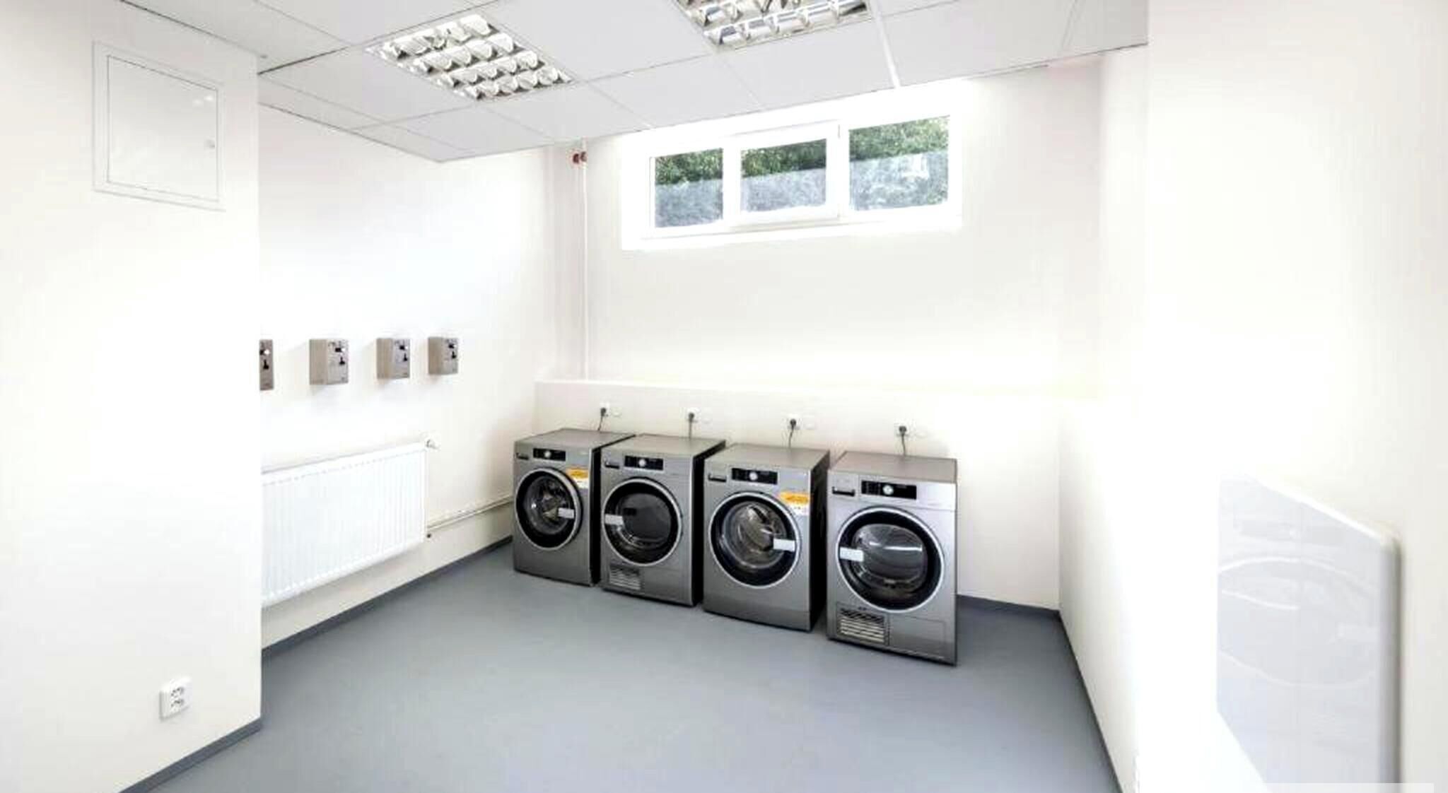 A laundry room with four front-loading washing machines, a window, and a radiator.