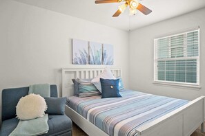 Un dormitorio con cama, una silla, ventilador de techo y ventana con persianas.