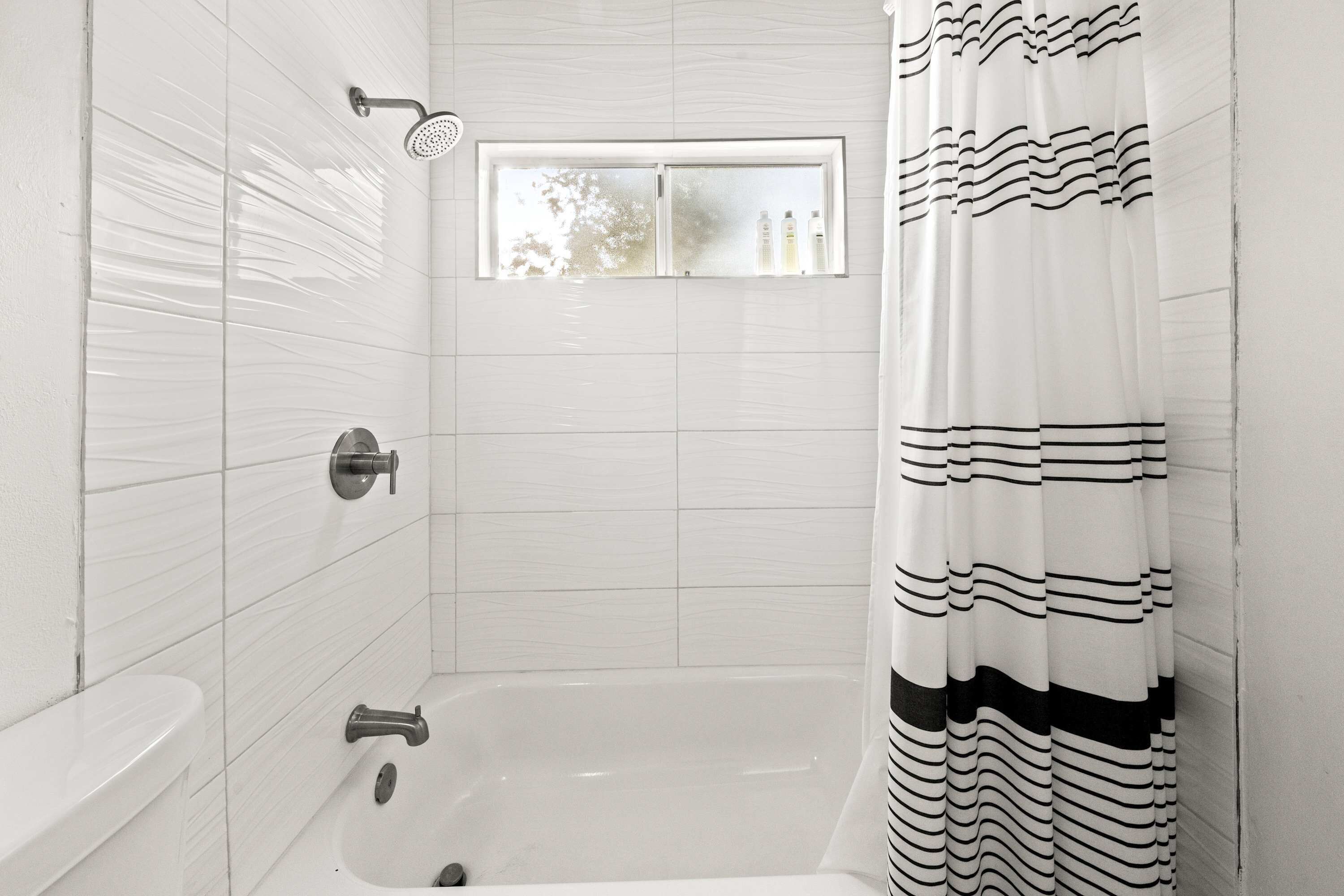 Une salle de bain avec des murs recouverts de carreaux blancs, une fenêtre, une douche et une baignoire.