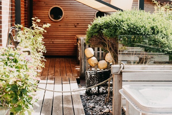 Une terrasse en bois avec une barrière en corde, un mur en bois doté de fenêtres rondes et un petit espace jardin avec des plantes et un banc en bois.