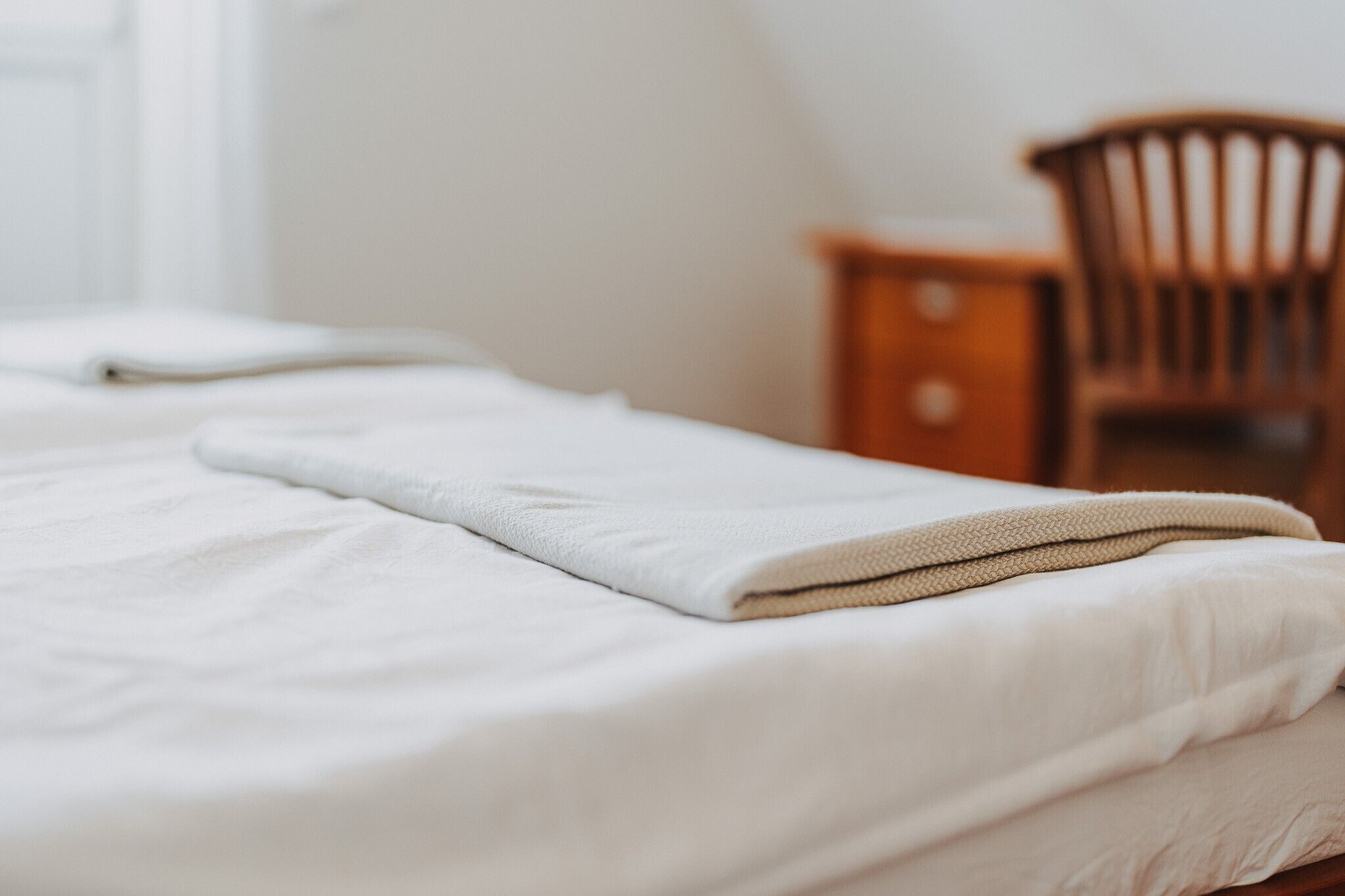 Ein ordentlich gemachtes Bett mit gefalteter Decke, ein Nachttisch aus Holz und ein Stuhl in einem Zimmer mit weißen Wänden.
