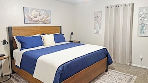Une chambre avec un lit en bois, une literie bleue, un mur blanc orné d’une œuvre d’art florale, un ventilateur de plafond et un tapis à motifs.