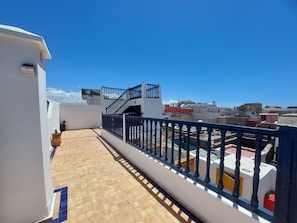 A rooftop terrace with a tiled floor, a black railing, and a view of the cityscape.
