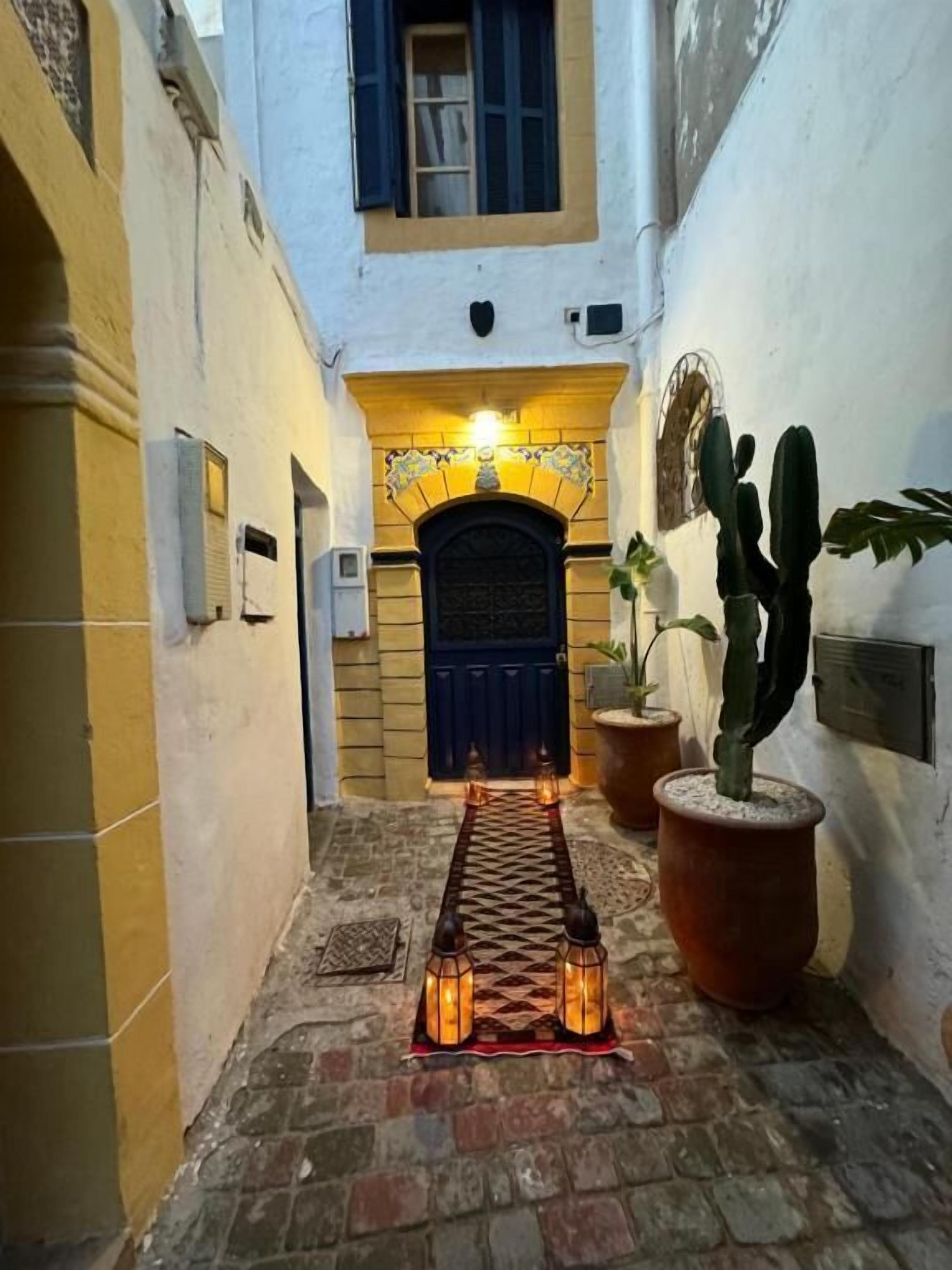 A narrow alleyway with a tiled floor, a dark door at the end, and potted plants along the walls.