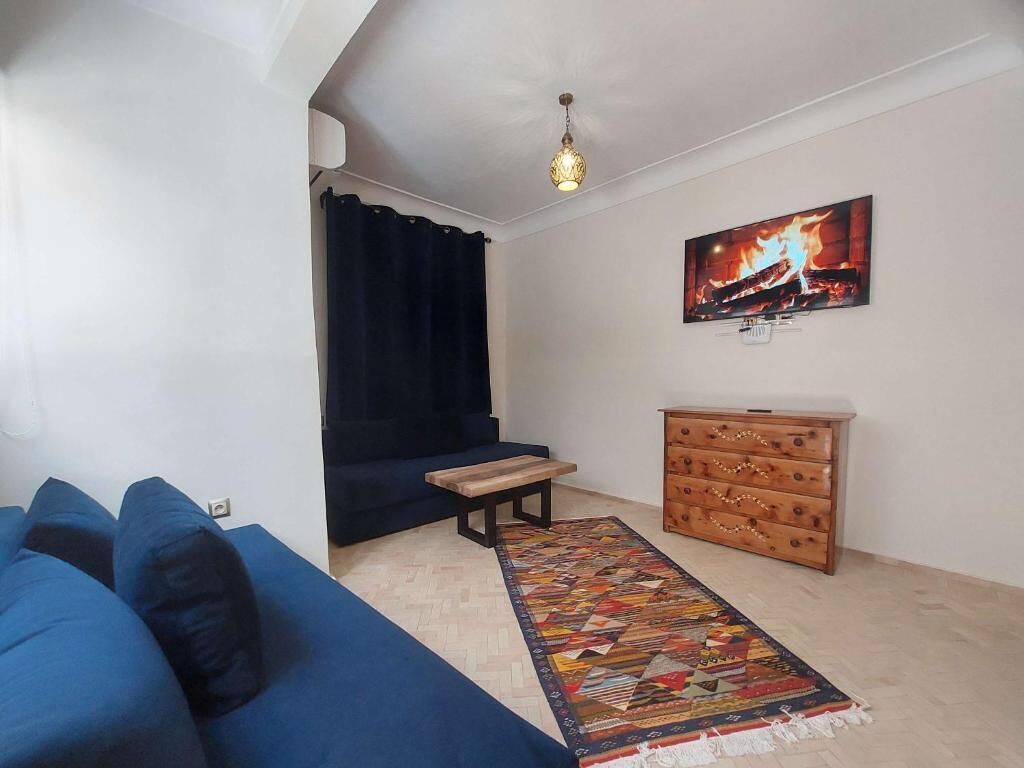 Ein Wohnzimmer mit einem blauen Sofa, einer Holzbank, einem gemusterten Teppich, einem Holzschrank und einem an der Wand befestigten Fernseher.