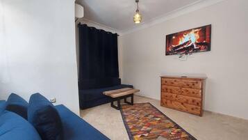 Ein Wohnzimmer mit einem blauen Sofa, einer Holzbank, einem gemusterten Teppich, einem Holzschrank und einem an der Wand befestigten Fernseher.