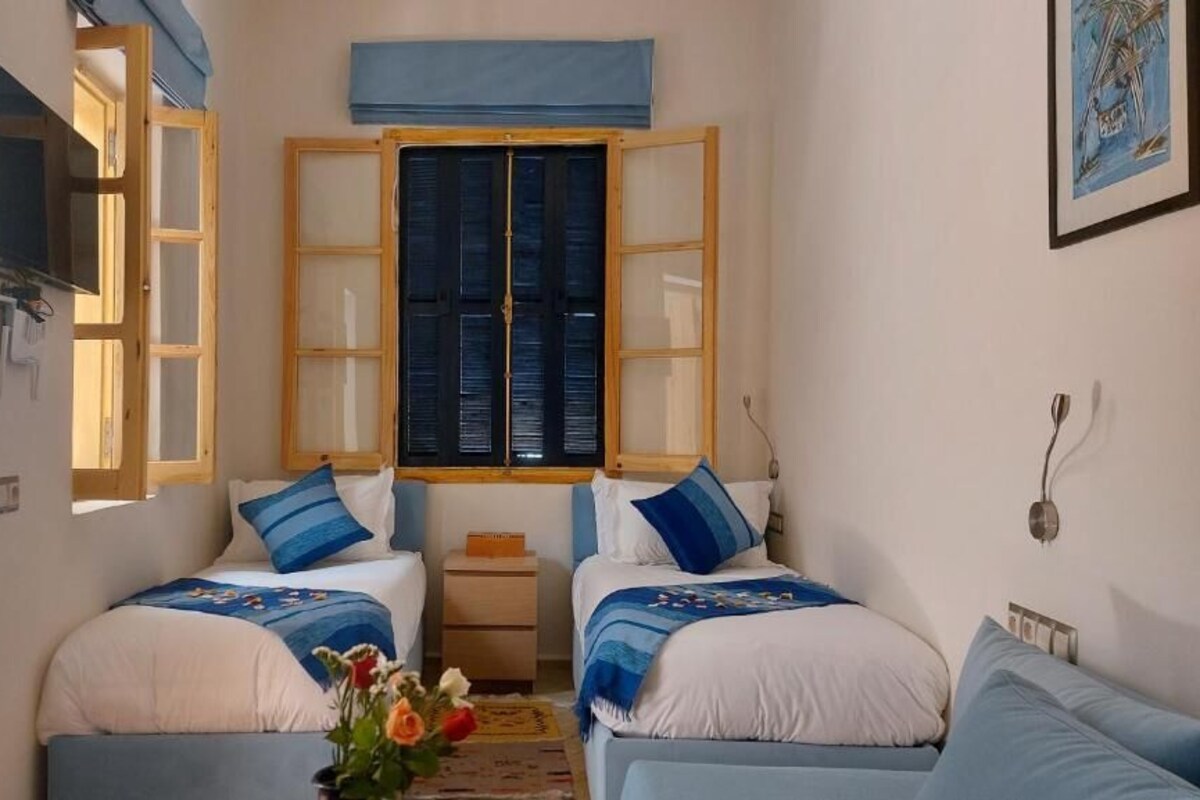 A small, cozy room with two beds, a television, a window with shutters, and a framed picture on the wall.