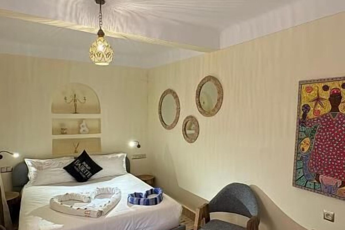 A bedroom with a bed, a wooden coffee table, two chairs, a colorful rug, and a ceiling light fixture.