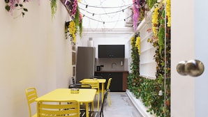 A narrow corridor with yellow tables and chairs, a kitchen area with a sink and stove, and walls adorned with hanging plants. - Sani Deluxe Suite Home (Athens)
