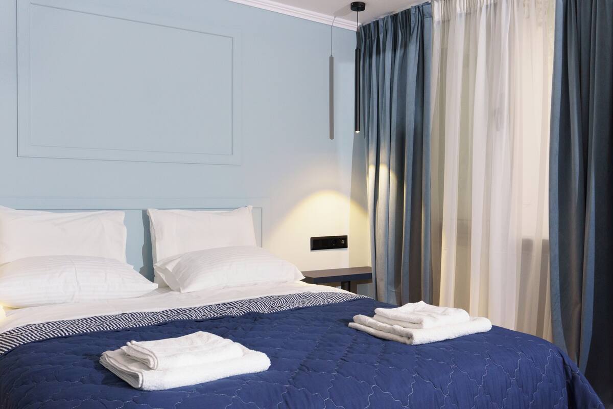 A neatly made bed with white pillows and a blue quilt, a bedside table with a lamp, and sheer curtains on a window.