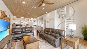 A spacious living room with a large leather sofa, a wooden coffee table, and a small side table with a lamp. A ceiling fan and recessed lighting are visible.