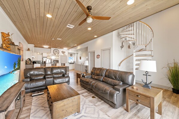 A spacious living room with a large leather sofa, a wooden coffee table, and a small side table with a lamp. A ceiling fan and recessed lighting are visible.