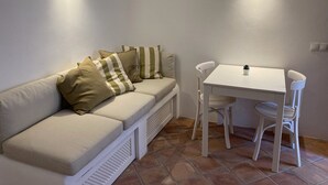 Um quarto pequeno e acolhedor, com um sofá branco, uma mesinha e duas cadeiras. Duas luzes fixadas na parede oferecem iluminação.