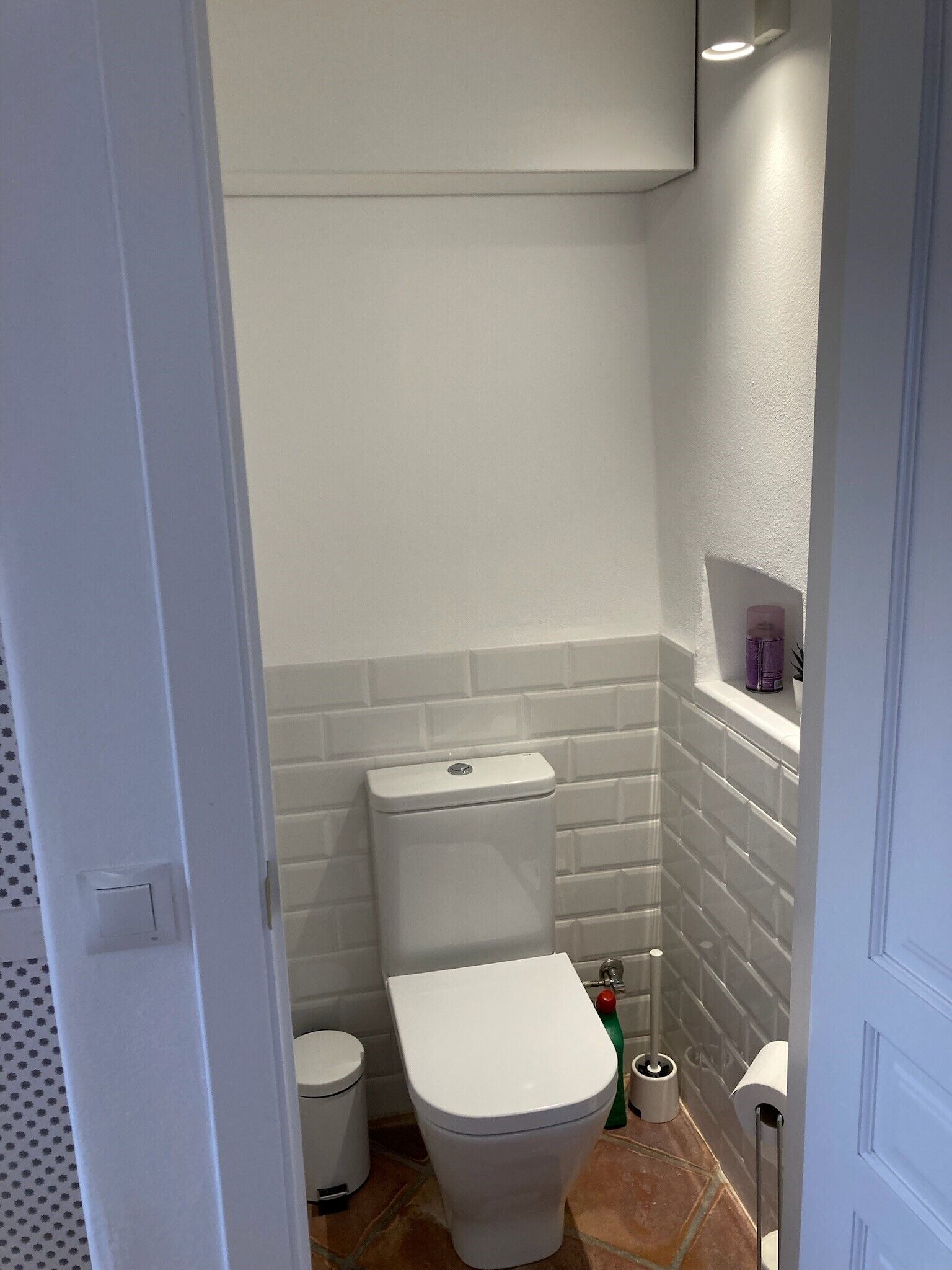 Une petite salle de bain compacte, avec des toilettes blanches, une petite poubelle et une brosse de nettoyage verte.