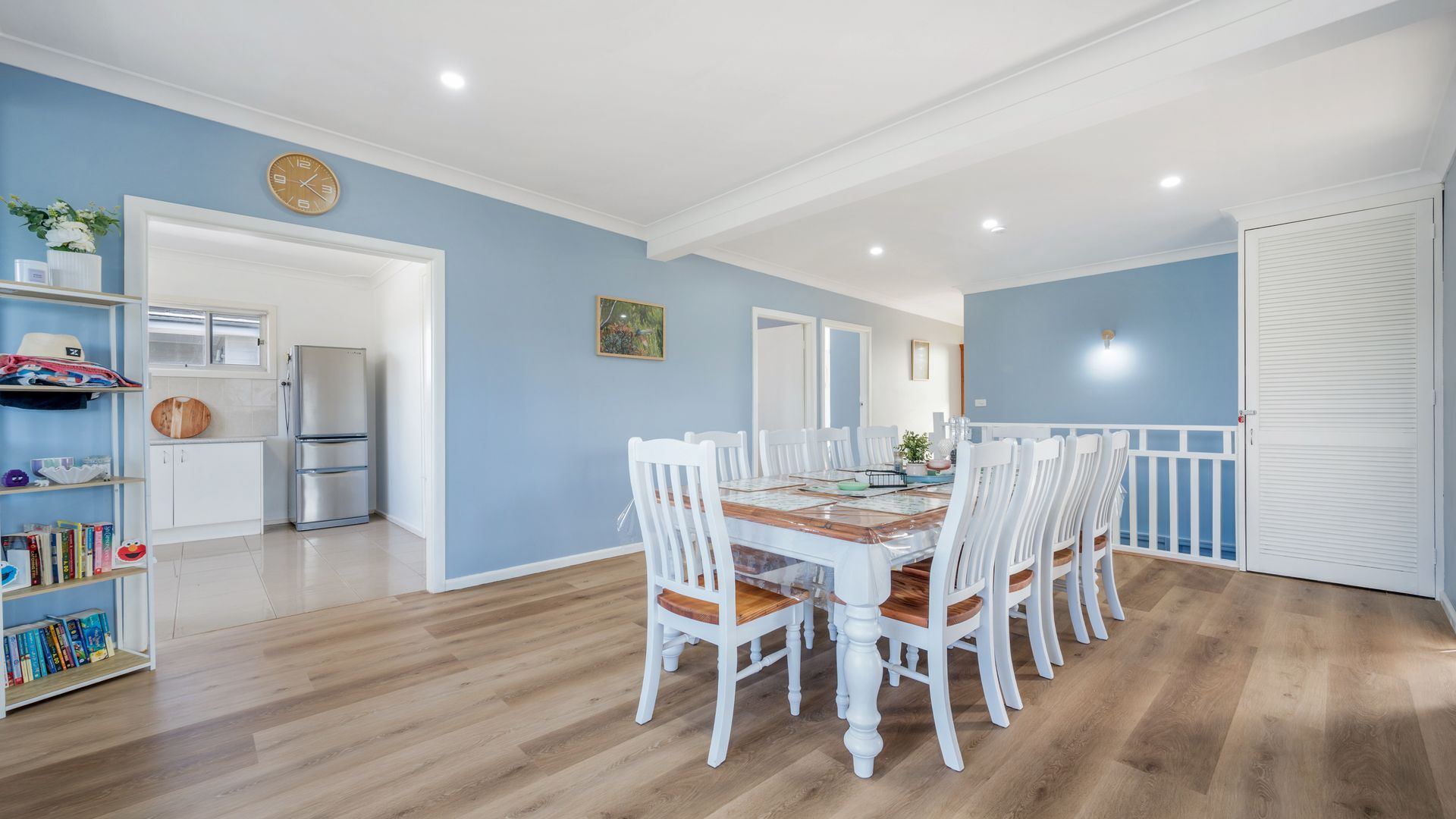 A dining area with a long table and white chairs, a kitchen with a refrigerator, and a blue wall with a clock.