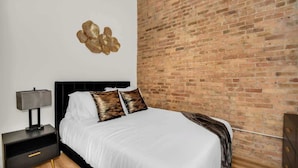 A bedroom with a brick wall, a bed with white bedding, two decorative pillows, a nightstand with a lamp, and a red square on the wall. - River North Rooftop With In and Out Parking 1 (Chicago)