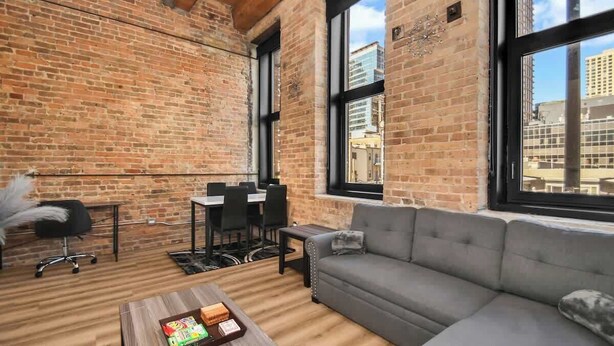 A spacious room with exposed brick walls, a large window with a city view, a gray sofa, a dining table with chairs, and a small side table. - River North Rooftop With In and Out Parking 1 (Chicago)