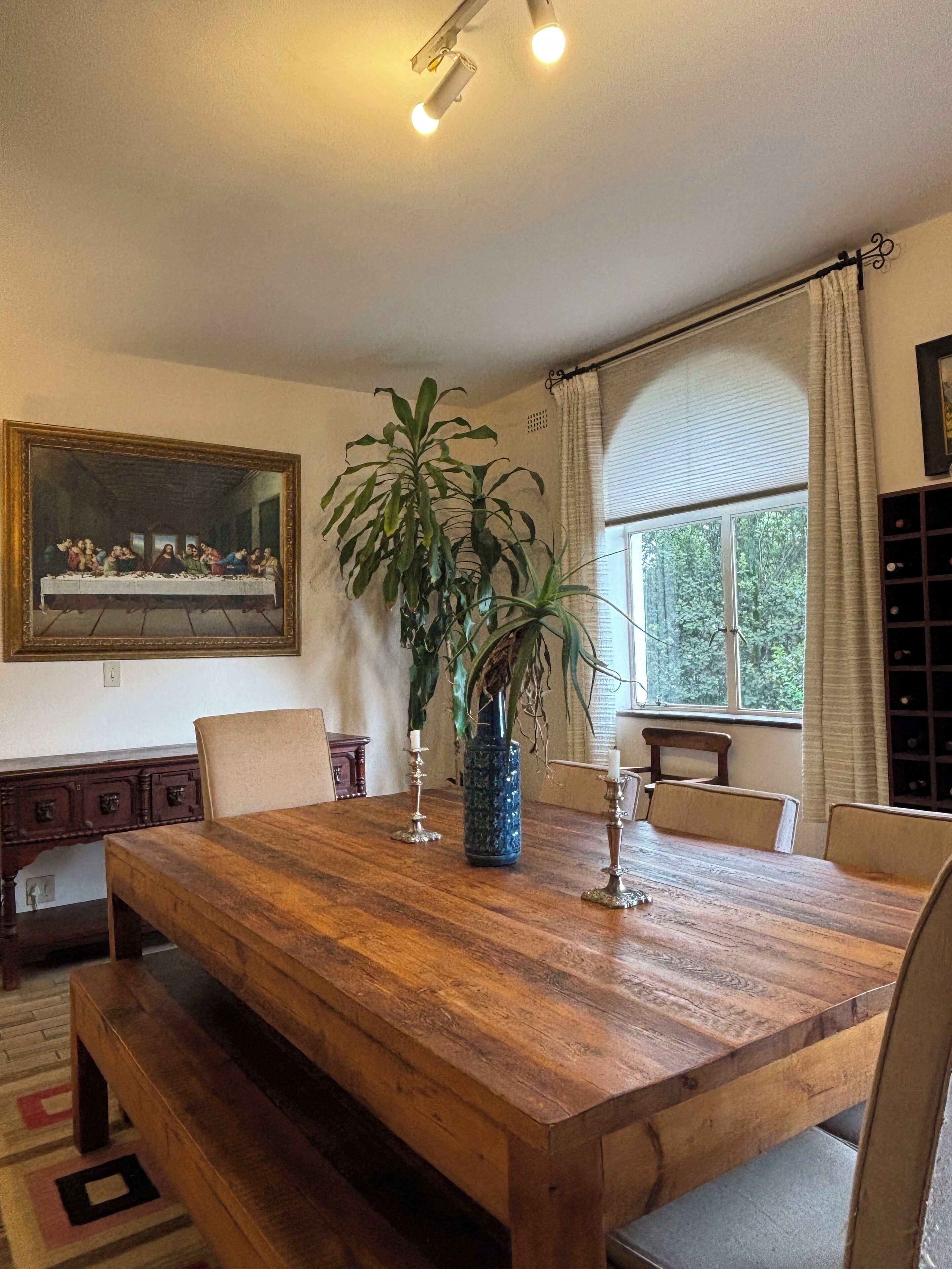 A dining room with a wooden table, chairs, a large potted plant, and a painting on the wall.