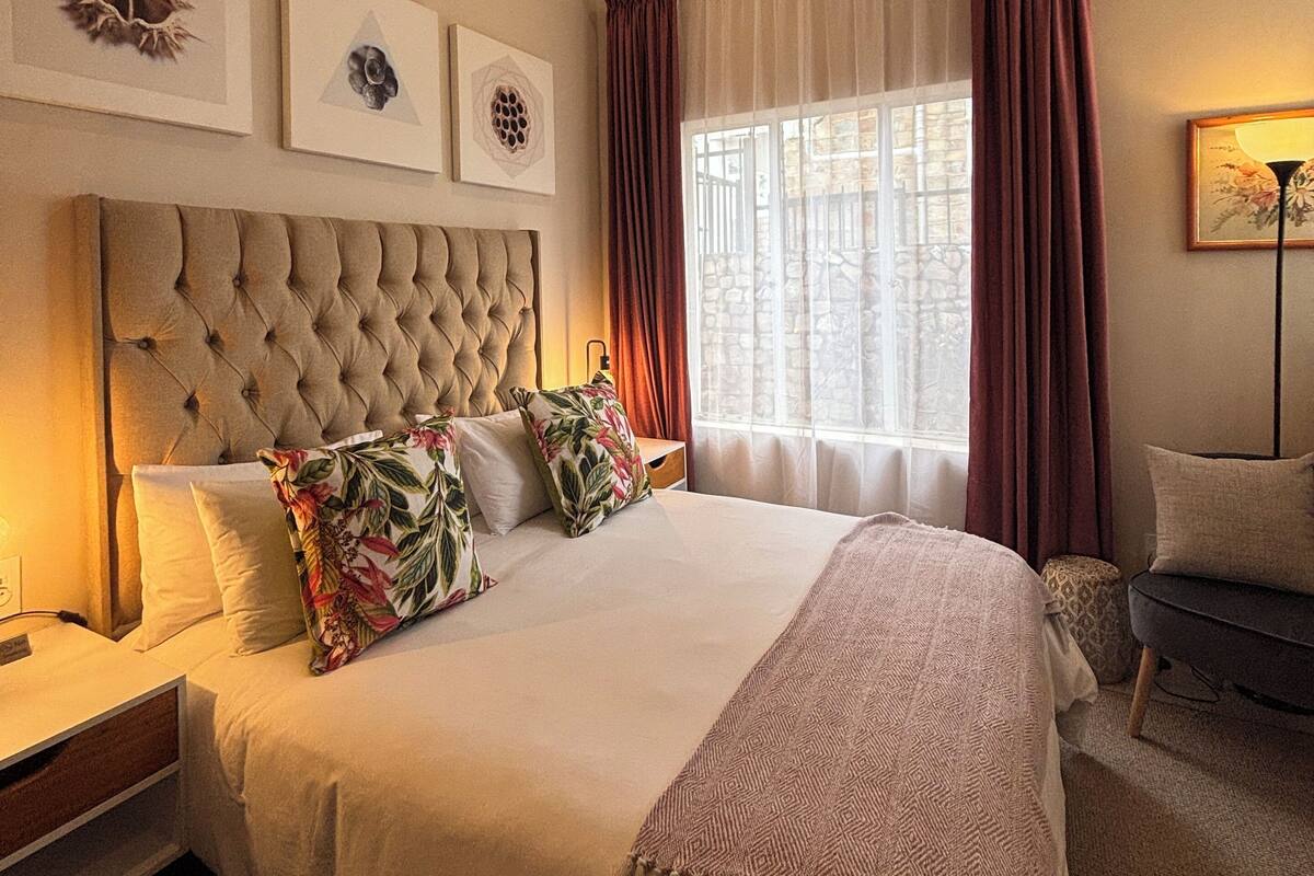 Un dormitorio luminoso con un cabecero capitonado, una cama grande con almohadas estampadas, una mesita de noche, una lámpara de pie y una ventana con cortinas.