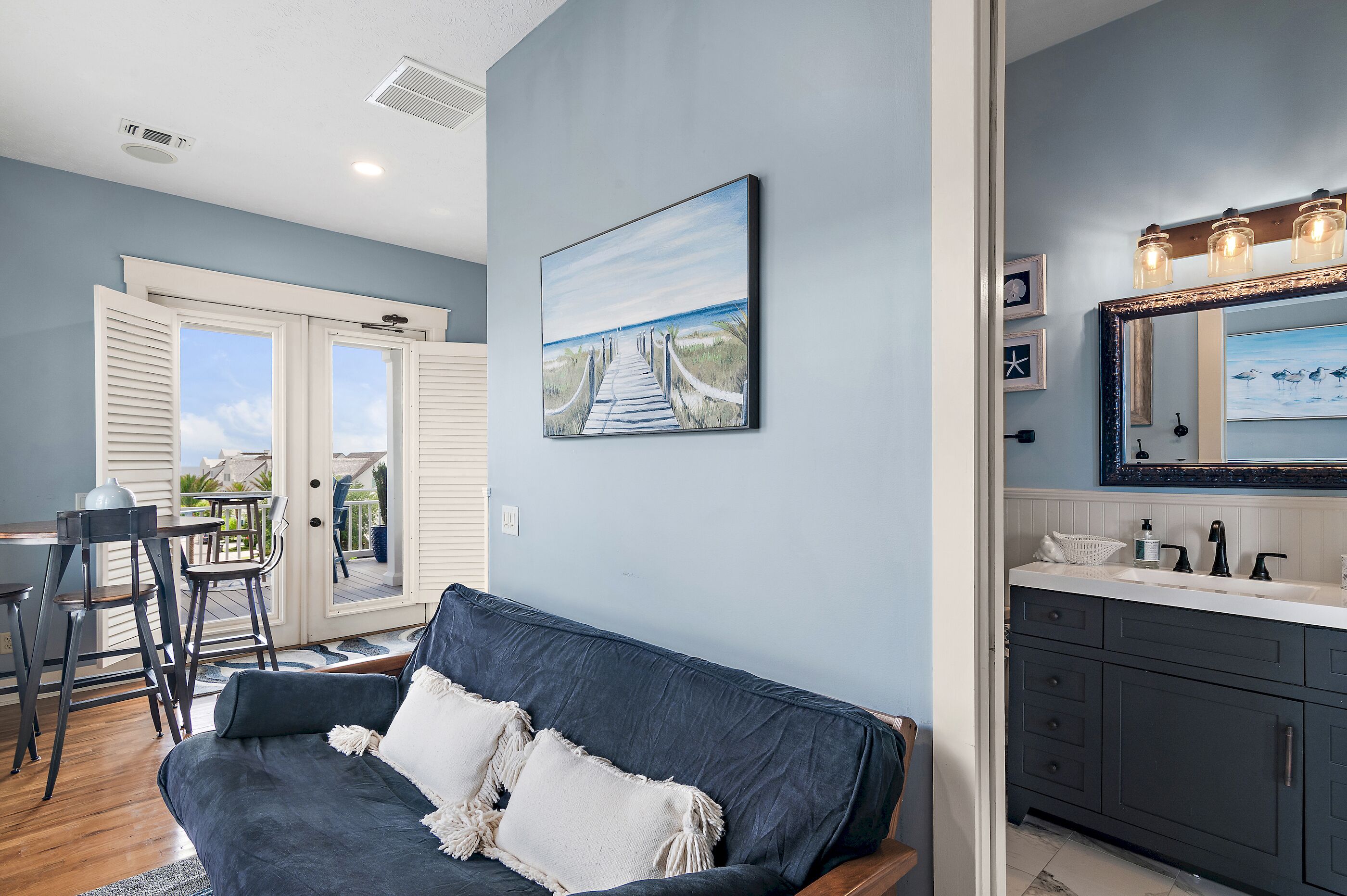Una sala moderna con un sofá azul oscuro, un cuadro de un camino de playa y una cocina con un espejo grande y lámparas colgantes.