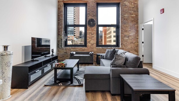 A modern living room with a brick wall, large windows, a flat-screen TV, a sofa, a coffee table, and a side table. - River North Rooftop With In and Out Parking 6 (Chicago)