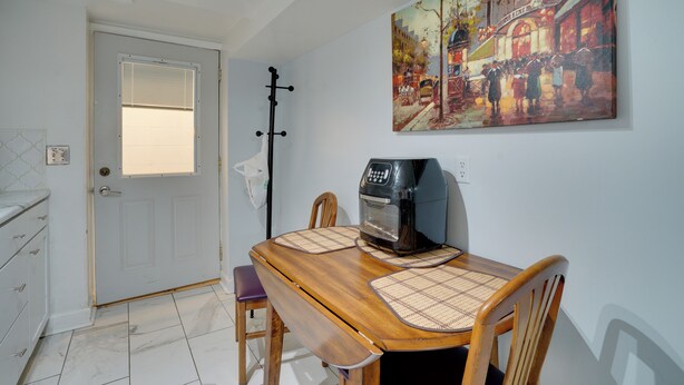 Un comedor con mesa de madera, sillas y una cafetera. Se observa una puerta con ventana y un cuadro en la pared.