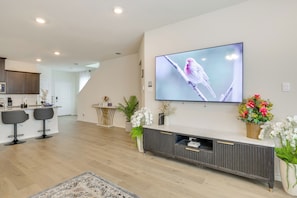 A modern living room with a flat-screen TV, a dark wooden entertainment unit, and a kitchen area in the background.