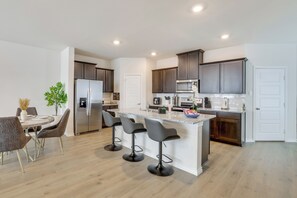A modern kitchen with a central island, dark cabinets, stainless steel appliances, and a dining area with chairs and a table. - 12 Mi to Dtwn Mckinney: Modern Gem w/ Patio & Yard (Anna)
