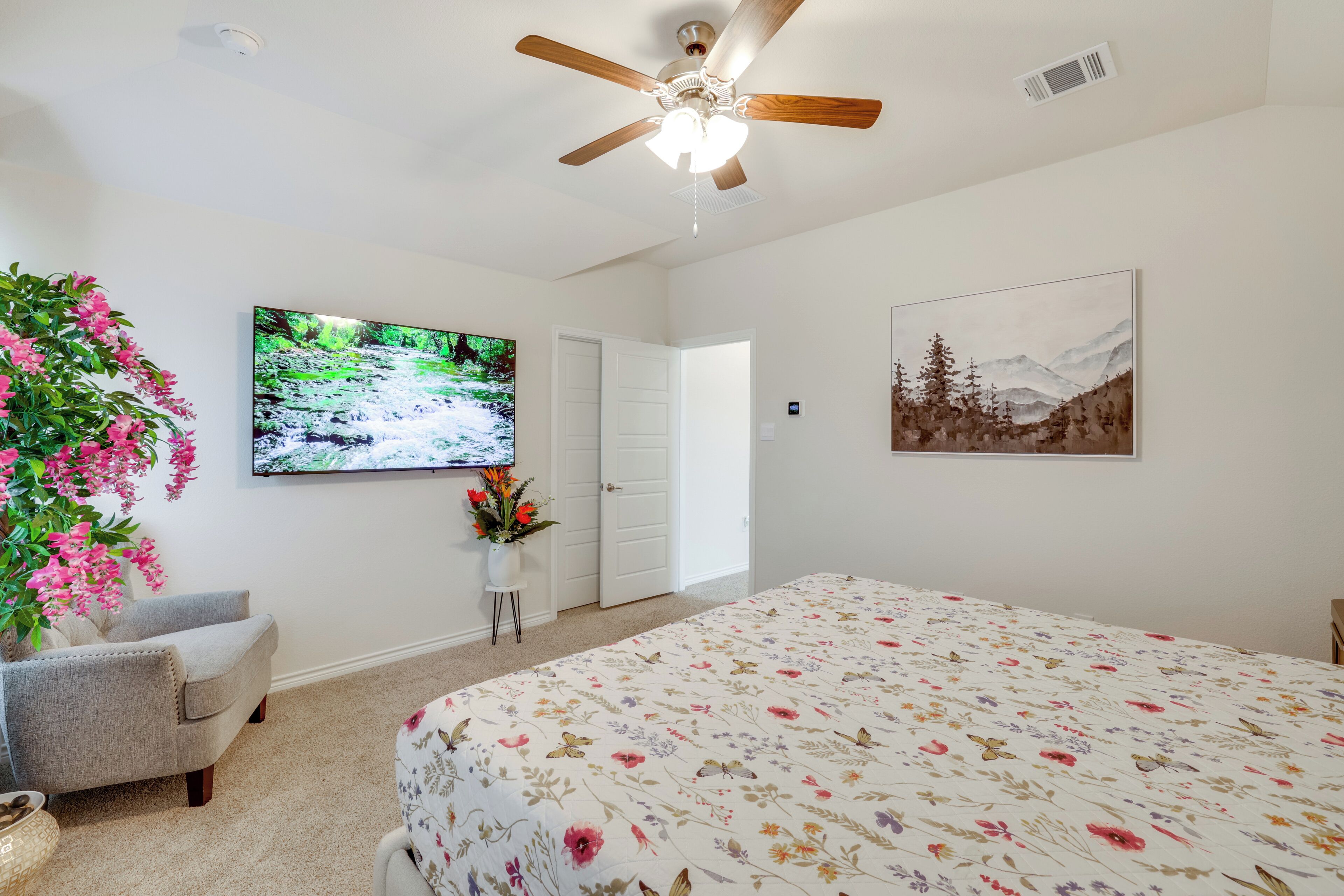A bedroom with a large bed, a ceiling fan, a TV on the wall, a door, and a painting.